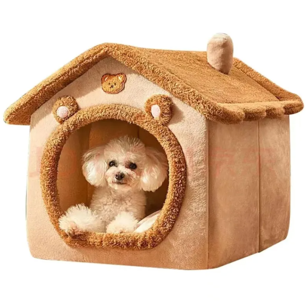 Small white dog sitting inside a tan house-shaped pet bed with a round, trimmed entrance.