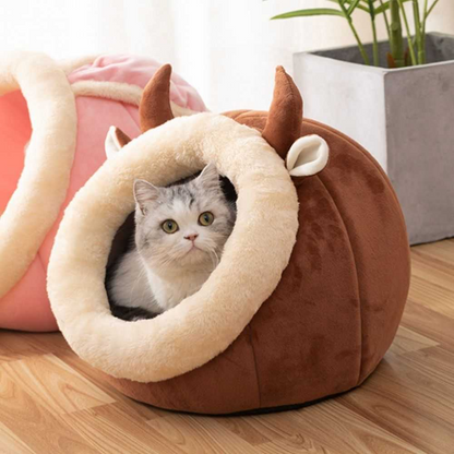 Brown rounded cat bed with a cream oval entrance and a gray cat sitting inside.