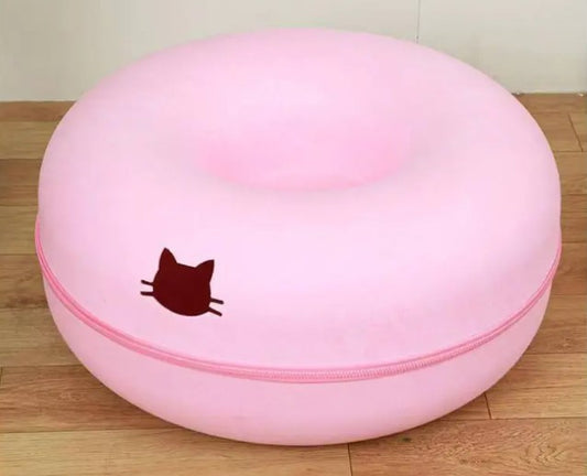 Pink round cat tunnel with a recessed center, pink trim, and a dark cat-head emblem on the front, photographed on a wooden floor.