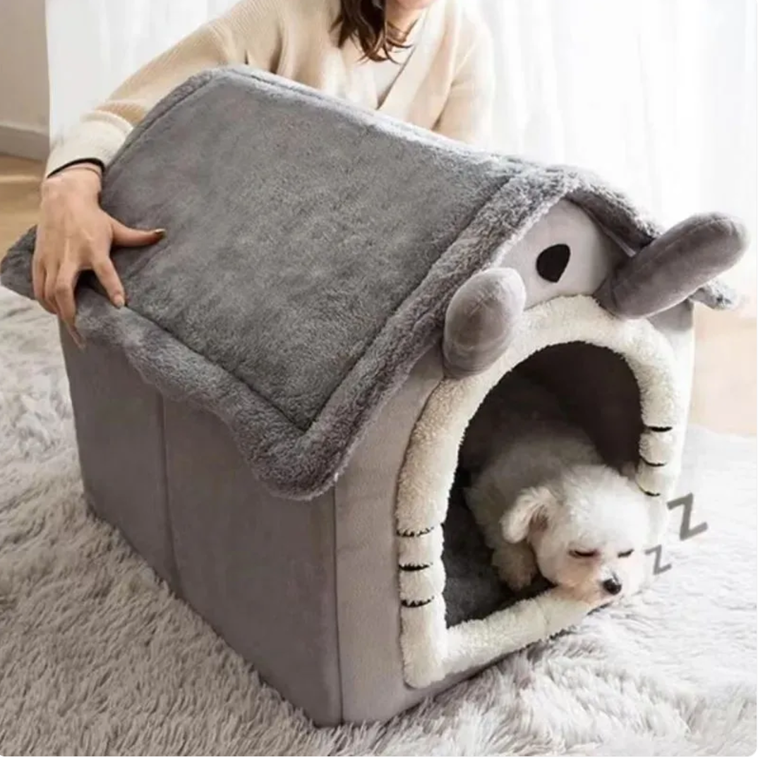 A person holds the roof of a gray house-shaped pet bed while a small white dog sits at the entrance.