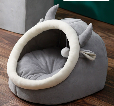 Gray enclosed cat nest bed with a plush white oval entrance, pointed ears and a dangling ball toy.