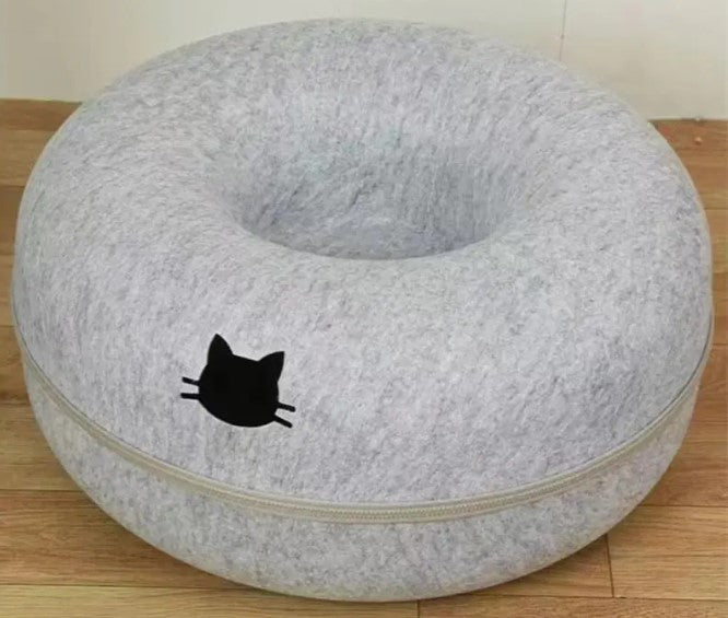 Light gray round cat tunnel with a recessed center, pale trim, and a black cat-head emblem on the front, photographed on a wooden floor.