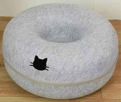 Light gray round cat tunnel with a recessed center, pale trim, and a black cat-head emblem on the front, photographed on a wooden floor.