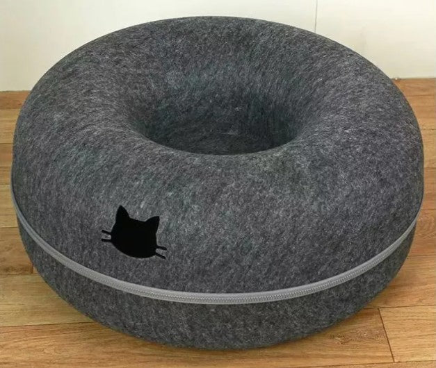 Dark gray round cat tunnel with a recessed center, pale trim, and a black cat-head emblem on the front, photographed on a wooden floor.